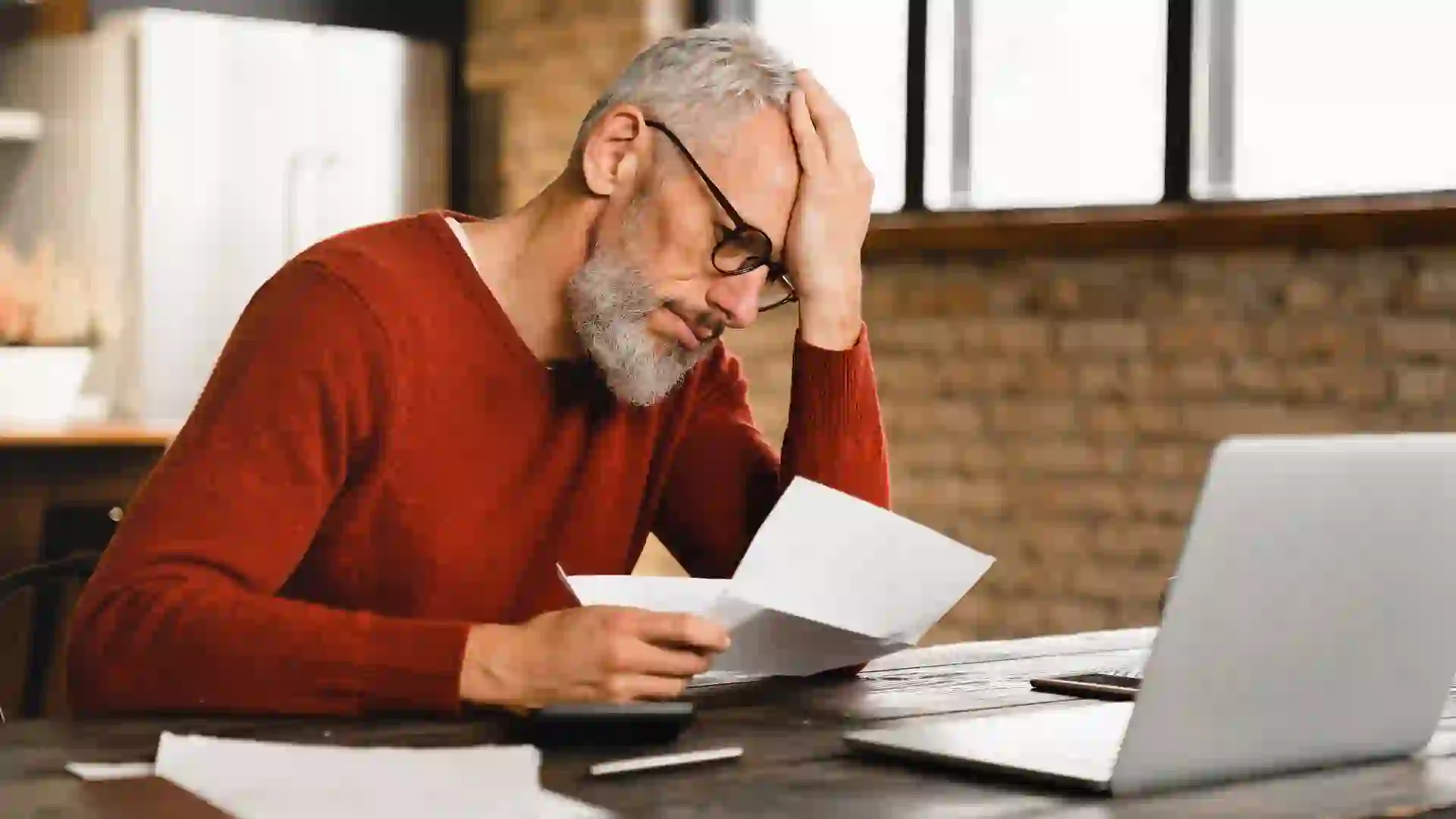 A man holds his head while reading a bill. Financial stress frustration over benefits, budget and other money worries.