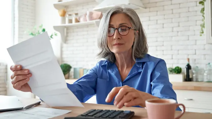 Senior mature business woman holding paper bill using calculator, old lady managing account finance, calculating money budget tax, planning banking loan debt pension payment sit at home kitchen table.