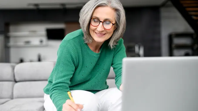 Confident mature woman using laptop computer for remote work, watching webinar and taking notes sitting at home.