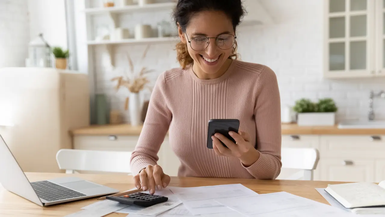 A happy woman smiles as she looks at her phone in her hand while sitting down at her desk in the kitchen
