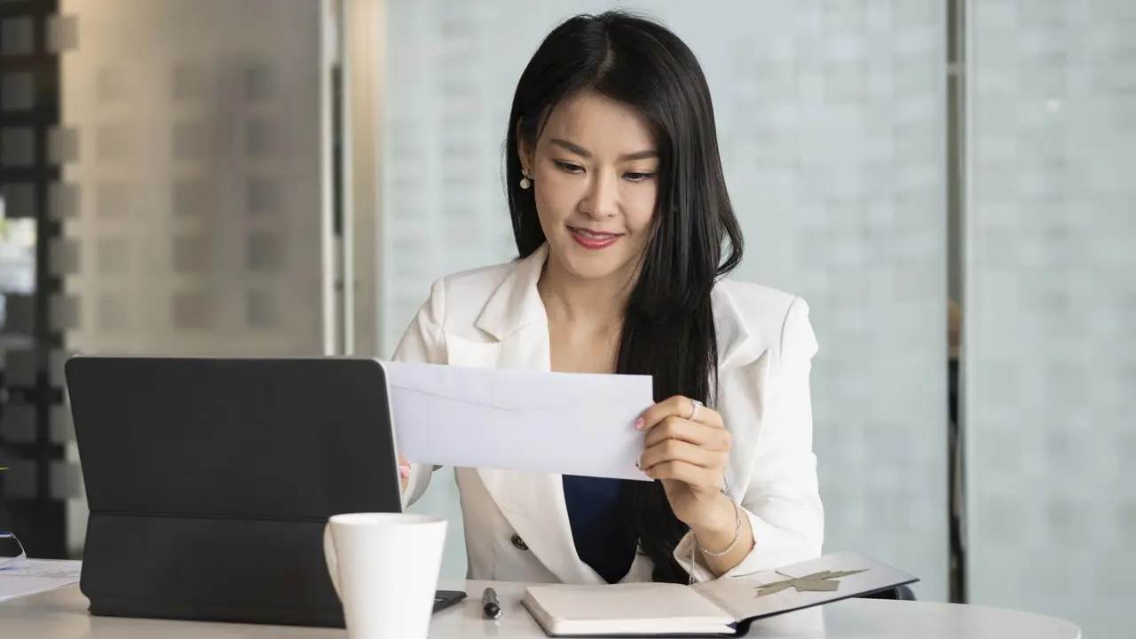 Smiling businesswoman sitting at her office desk holding envelope.