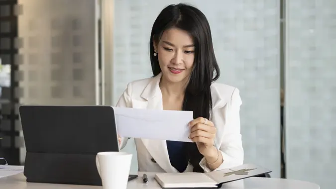 A young woman smiles while sitting at a desk and holding an envelope.