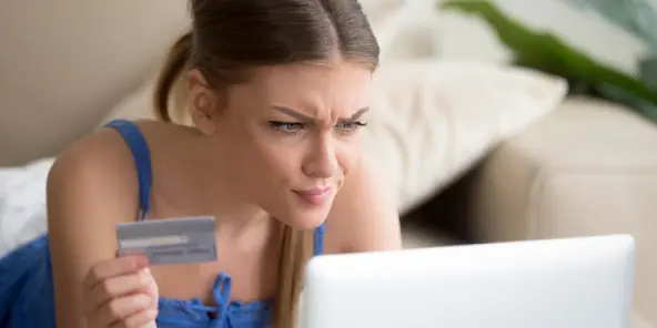 A skeptical woman looking confused at her laptop while holding her credit card, scrutinizing a bill or her credit report.