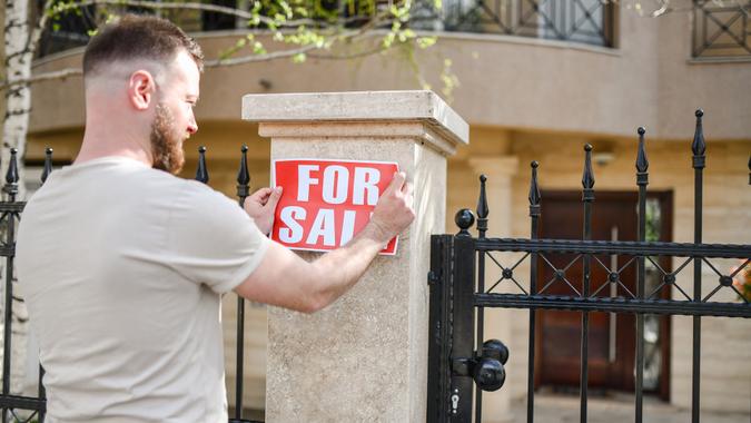 man hanging for sale sign home_iStock-1394994878 Bearded Male Declaring House For Sale stock photo