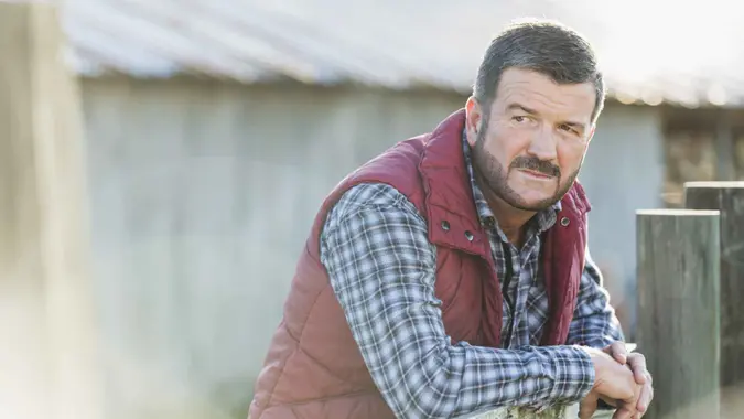A farmer or rancher, mature man in his 50s, standing outside a barn, with his arms leaning on a wooden fence.