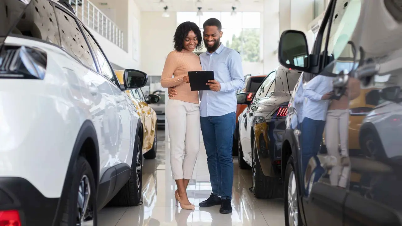 Young black couple choosing car in modern dealership center, holding clipboard and checking vehicle specs in catalog, happy african american spouses buying automobile in showroom.