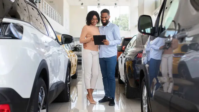 Young Black Couple Choosing Car In Modern Dealership Center, Holding Clipboard And Checking Vehicle Characteristics In Catalog, Happy African American Spouses Buying Automobile In Showroom.