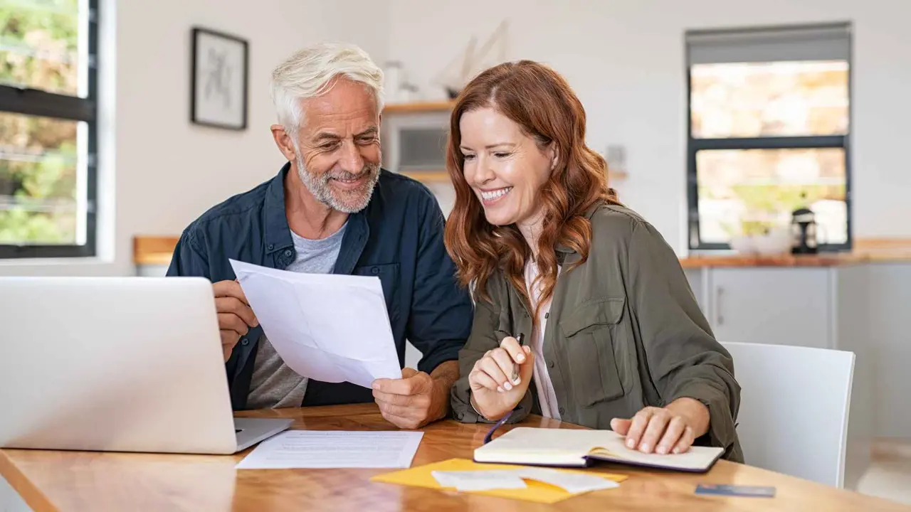 A happy couple sits at the table and smiles while sitting at their table and looking at paperwork.