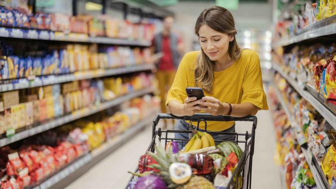 Purchasing Goods with Smartphone at Grocery Store Female customer shopping with smartphone checklist, taking products from shelf at the shop.