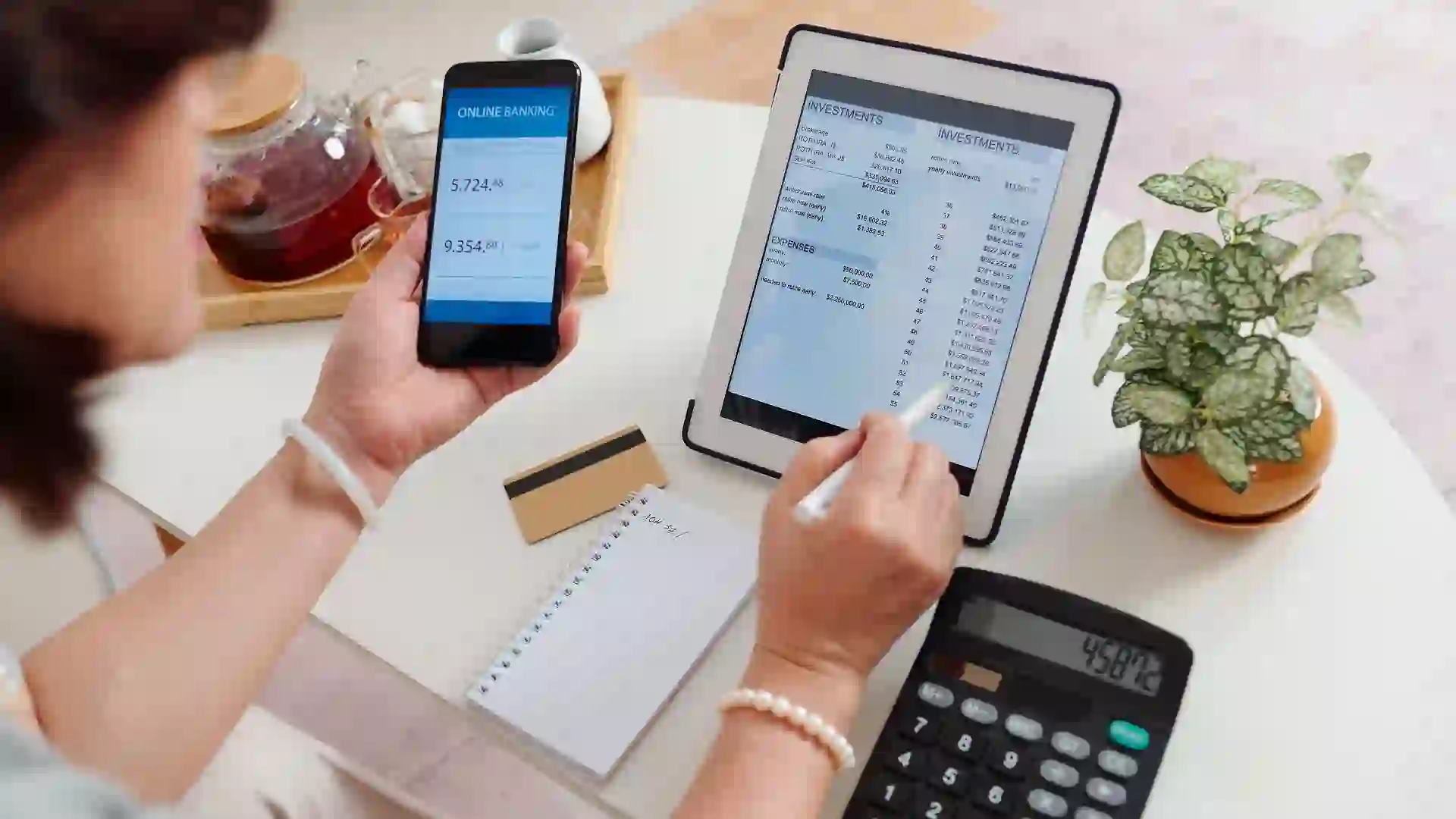 Woman checking brokerage account on a phone and tablet with a calculator, note paper and a credit card on her desk