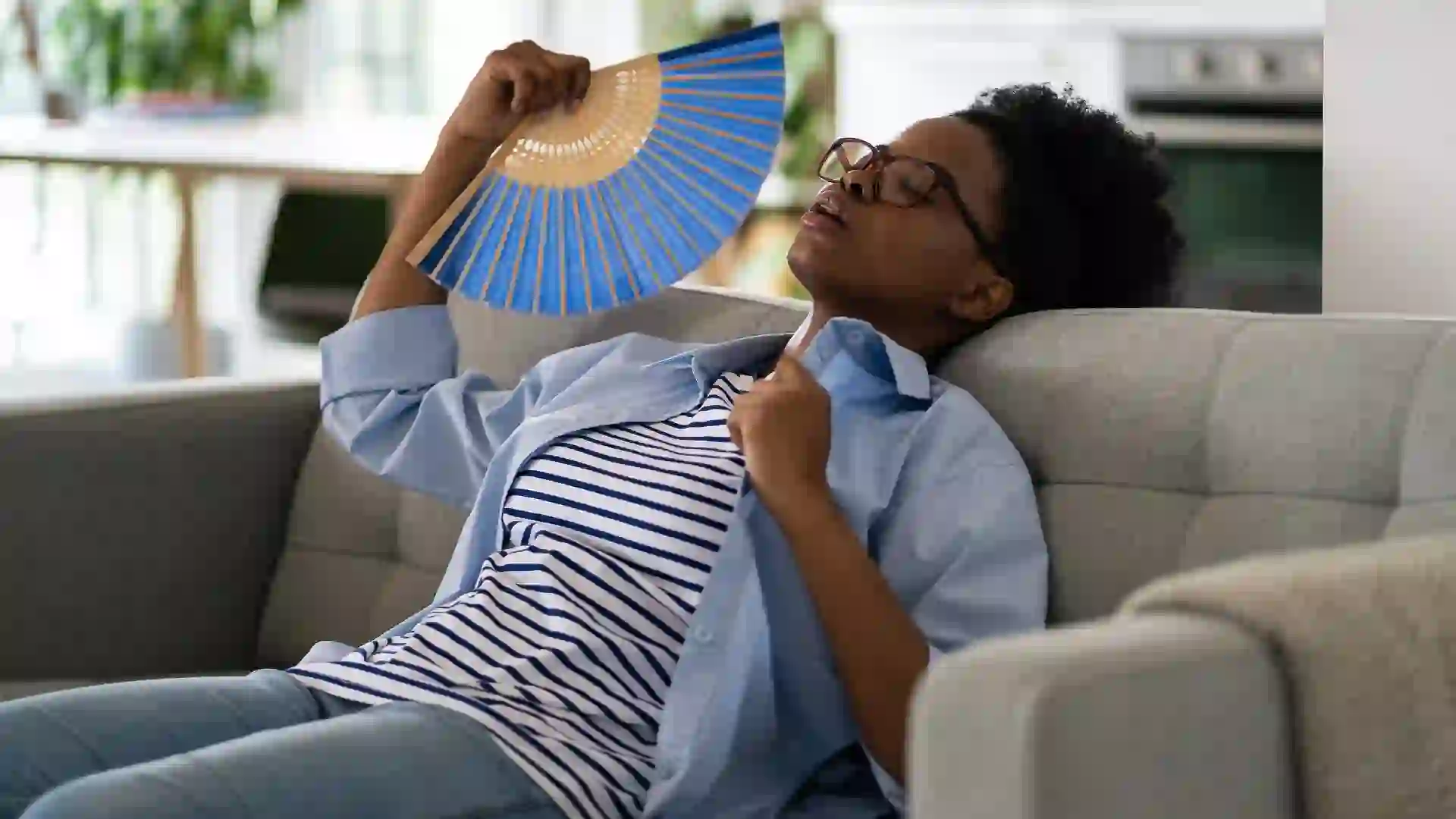 Tired exhausted African American woman sits on sofa using paper fans suffering from stuffiness stock photo
