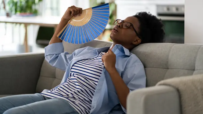 Tired exhausted African American woman sits on sofa using paper fans suffering from stuffiness stock photo