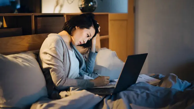 Busy young Asian woman sitting on bed, working from home using laptop and handling paperworks till late in the evening at home, looking stressful.
