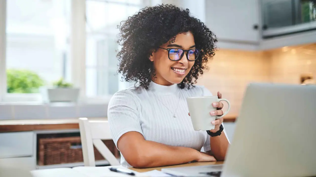 A woman smiles as she drinks coffee and works on her laptop.