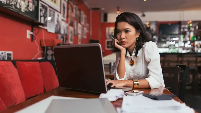 Pensive Asian bar owner using laptop to pay bills online.