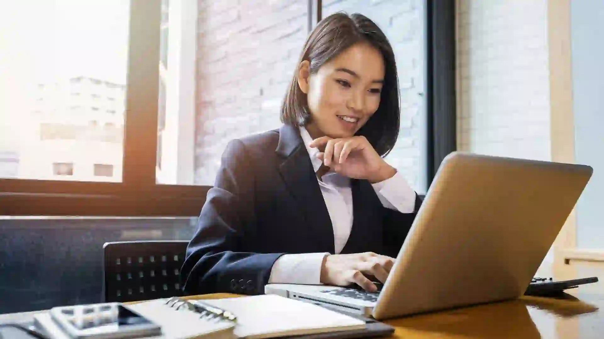 Shot of a woman sitting down in her work office smiling as she is looking at her open laptop.