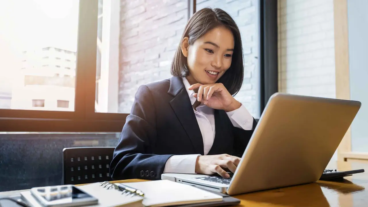 Shot of a woman sitting down in her work office smiling as she is looking at her open laptop.