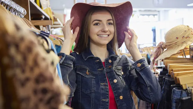 Young Woman Buying Used Sustainable Clothes From Second Hand Charity Shop Trying On Hat stock photo