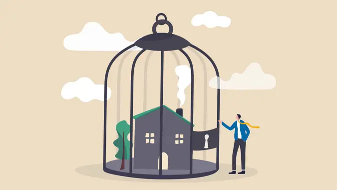 Mortgage payment problem, housing debt in economic crisis concept, worried house owner businessman standing with his house inside locked bird cage.