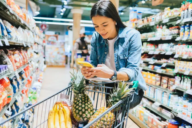 Happy young woman shopping for groceries in supermarket standing near her trolley using smartphone.
