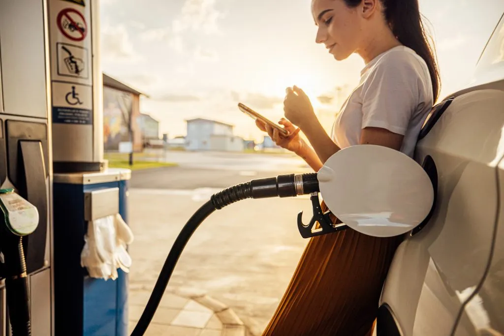 Beautiful young woman text messaging on smart phone while refueling gas tank at fuel pump.