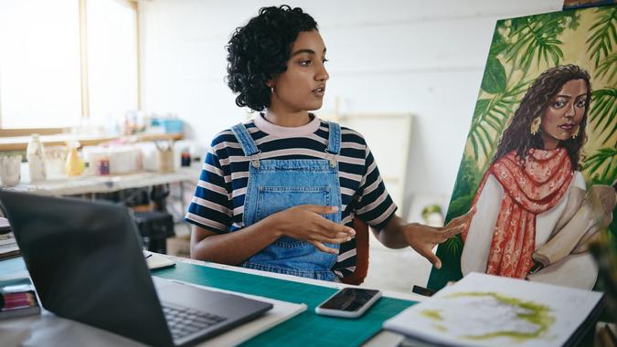 Art girl and laptop video call in studio for professional remote sale of painting on canvas online. Creative indian woman with focus and concentration selling artwork on the internet in workshop. stock photo