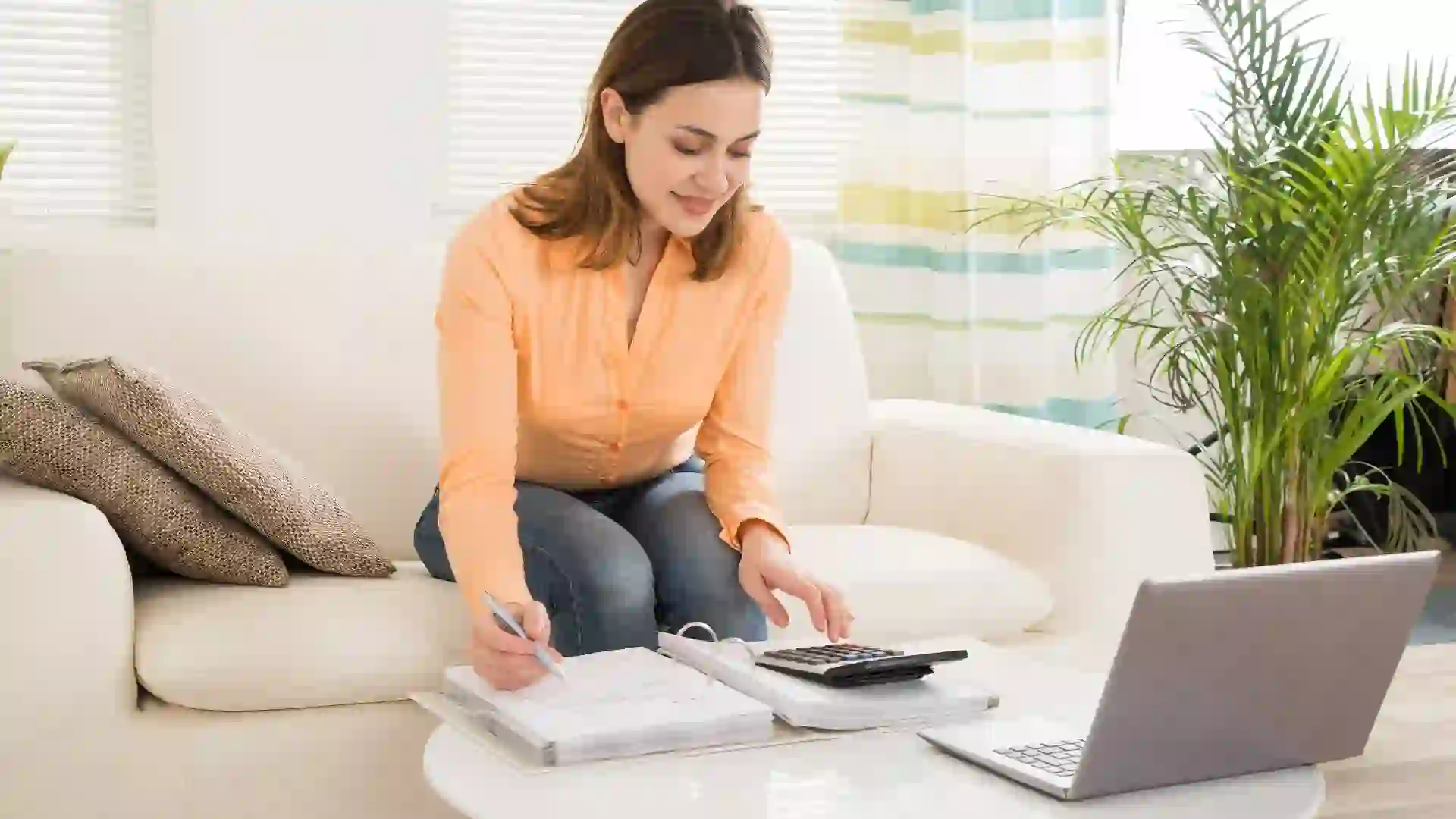A woman smiles as she calculates her bills and looks at her laptop to see how to save money.
