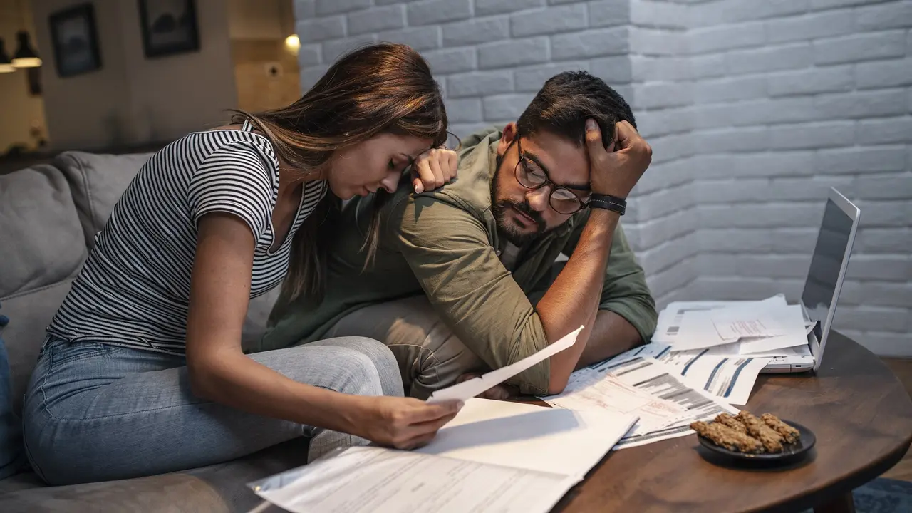 A couple sits at their kitchen table, visibly stressed and anxious as they go over a growing stack of unpaid bills.