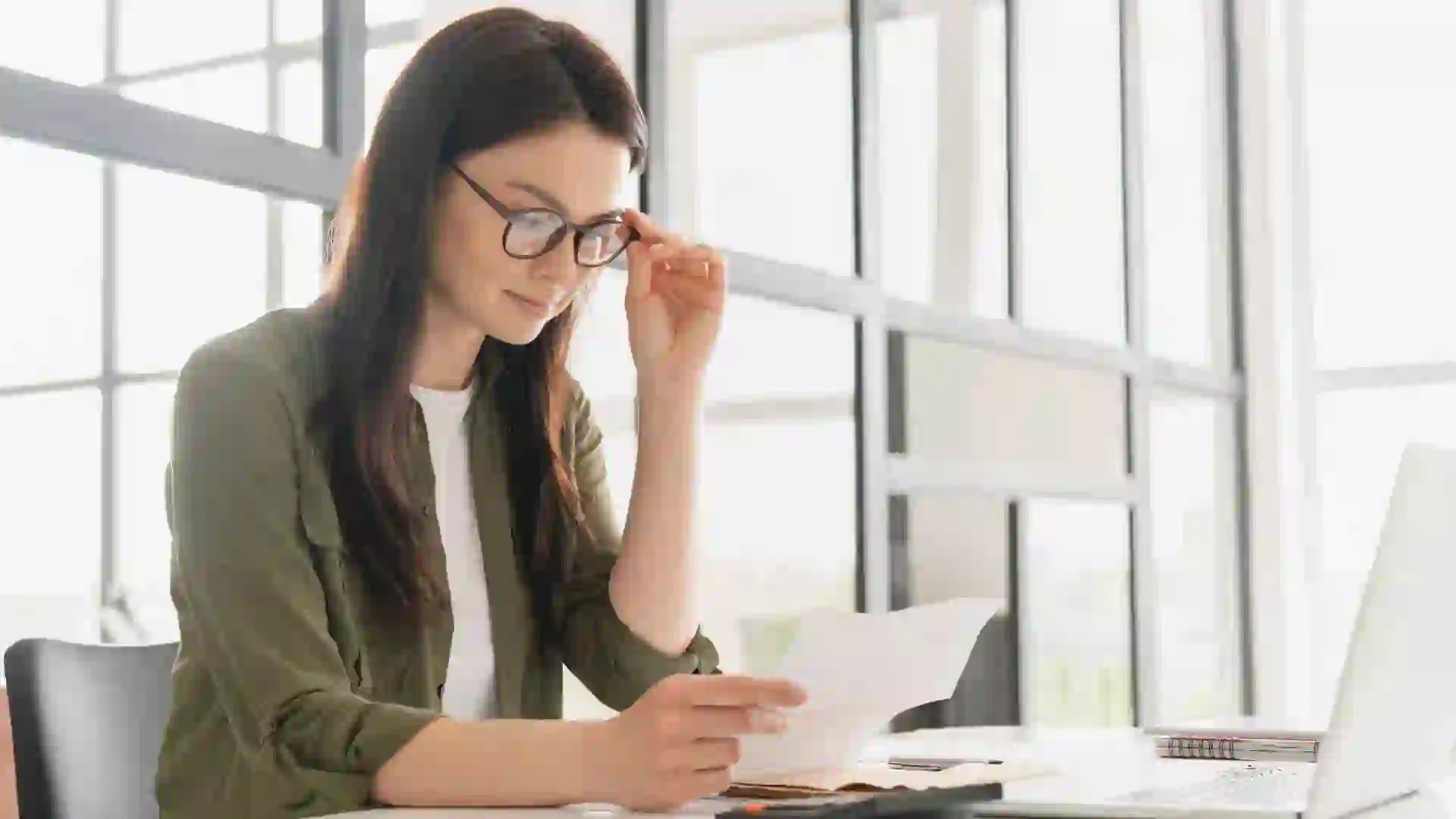Young businesswoman sitting in an office, looking over paperwork with her laptop in front of her.