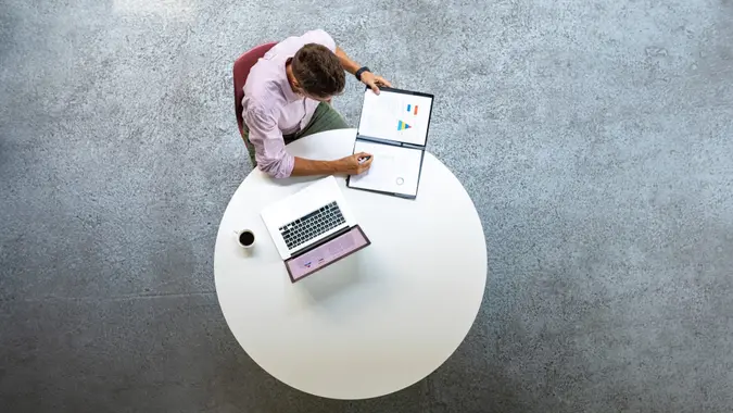 From above photo of businessman sitting at a round table and working with documents and a laptop.
