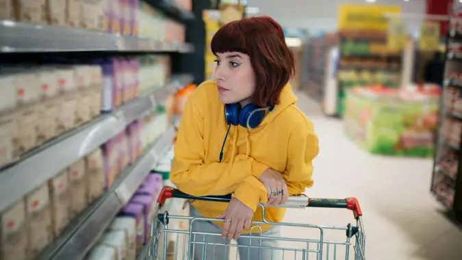Young woman with red hair doing groceries in a local supermarket, pushing a shopping cart and looking at the prices.