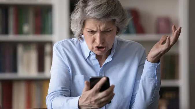 Angry older businesswoman sit at workplace staring at cellphone screen stock photo