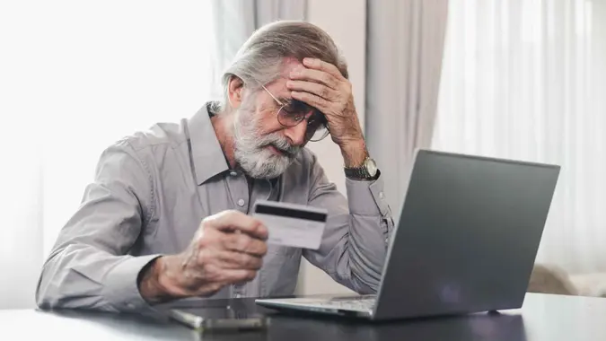 Upset senior elderly man holding credit card by laptop having trouble worry finance safety data or online payment security.