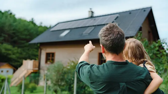 Rear view of dad holding her little girl in arms and showing at their house with installed solar panels. Alternative energy, saving resources and sustainable lifestyle concept. stock photo