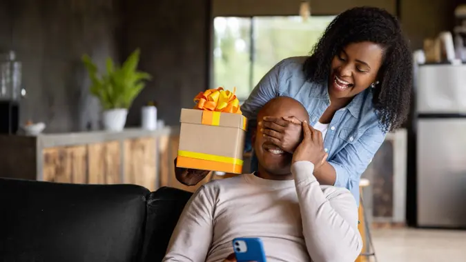 African American woman at home surprising her husband with a gift and covering his eyes.