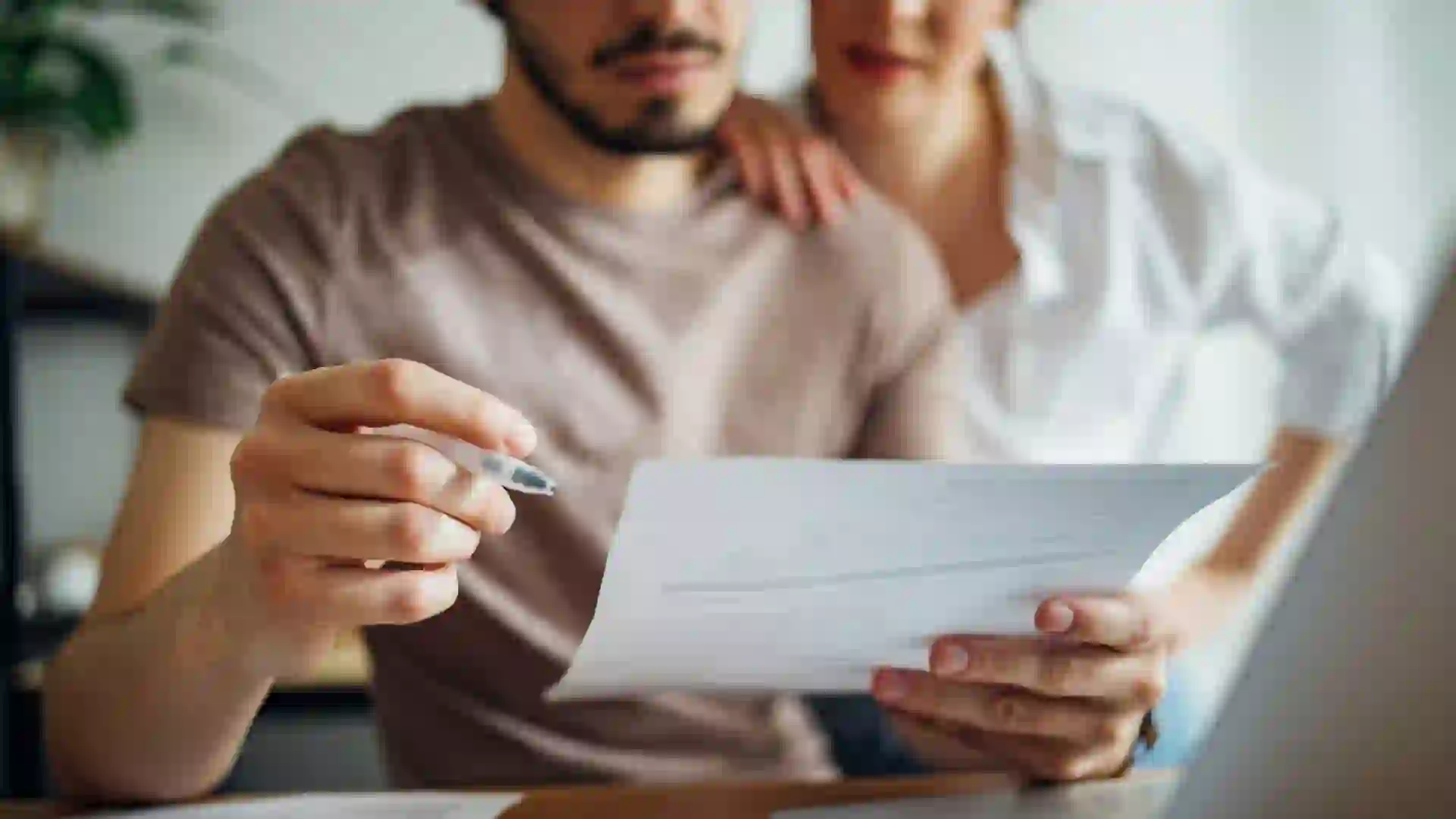 Close up photo of male hands holding bill and pen and female hands hugging him while they doing home finances together online on a laptop computer in the kitchen.