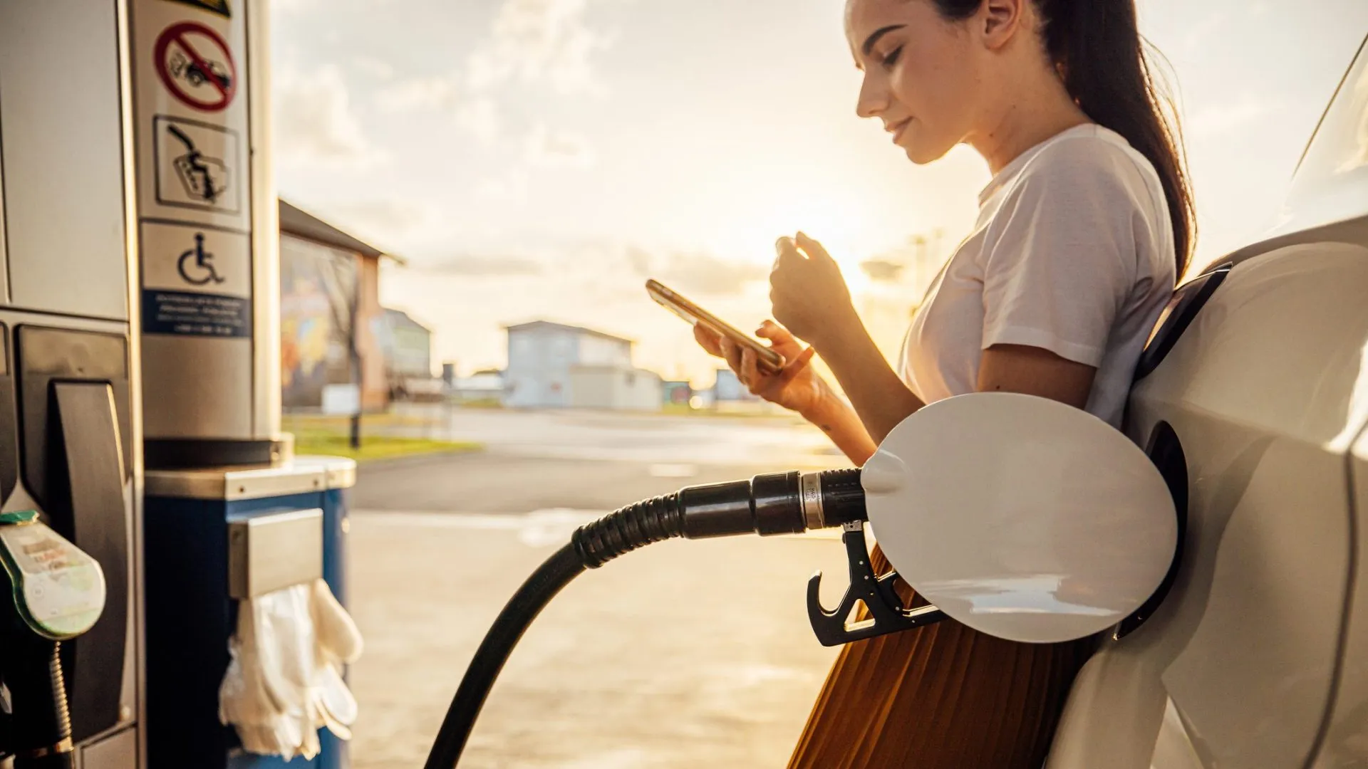 Beautiful young woman text messaging on smart phone while refueling gas tank at fuel pump.