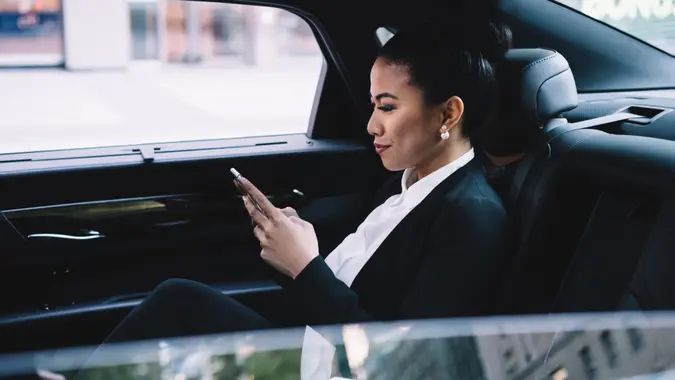 Confident adult Asian executive woman in formal clothes concentrating on screen and interacting with smartphone while sitting in luxurious automobile in city.