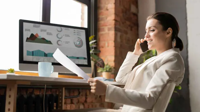 Happy young well-dressed accountant reading one of papers by desk and explaining data to colleague on the phone.