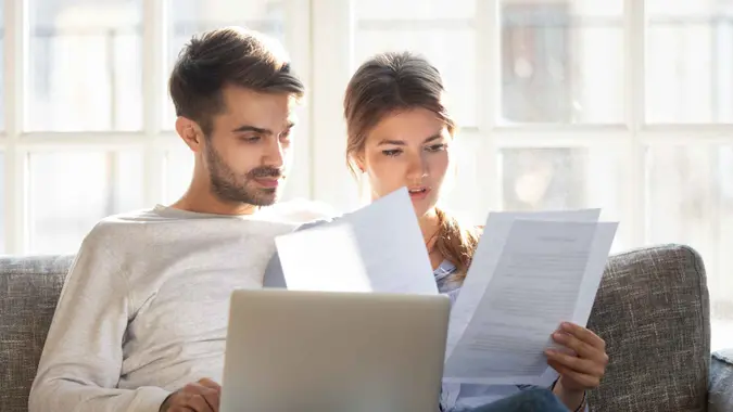 Millennial married couple sitting on couch with computer and documents, reading received formal letter from bank loan mortgage information, contract terms and conditions discuss papers working concept.
