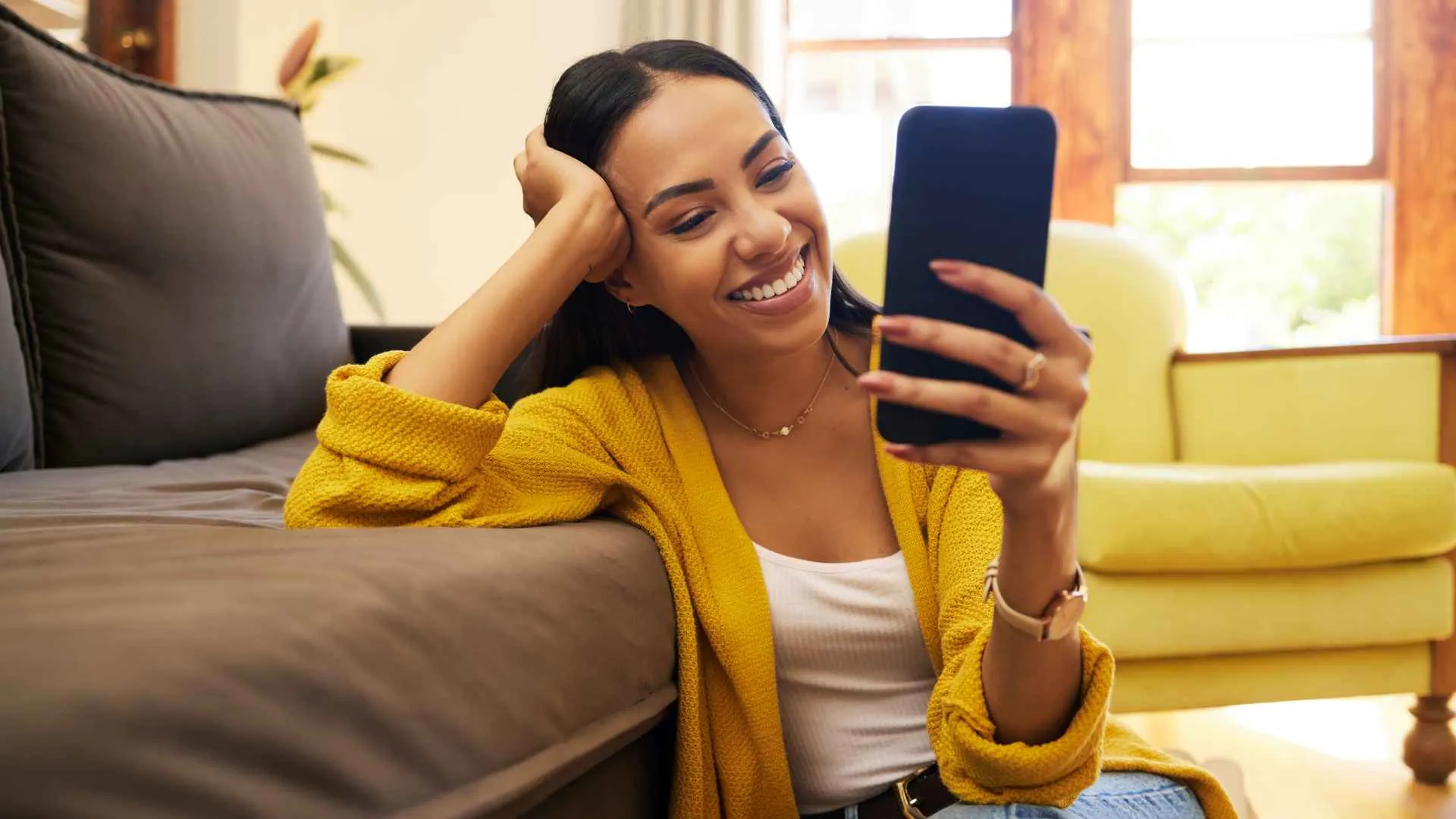 Woman smiling at her cellphone at home sitting on the floor against a sofa in a bright living room.