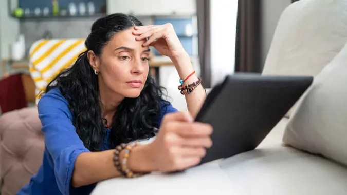 Pretty curly woman sitting on the sofa, holding a digital tablet, reading and looking concentrated and concern.