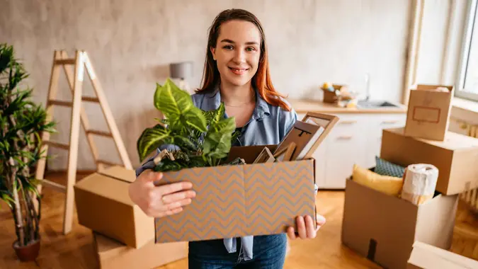 Portrait of beautiful young woman holding cardboard box with plants and picture frames in her new apartment.