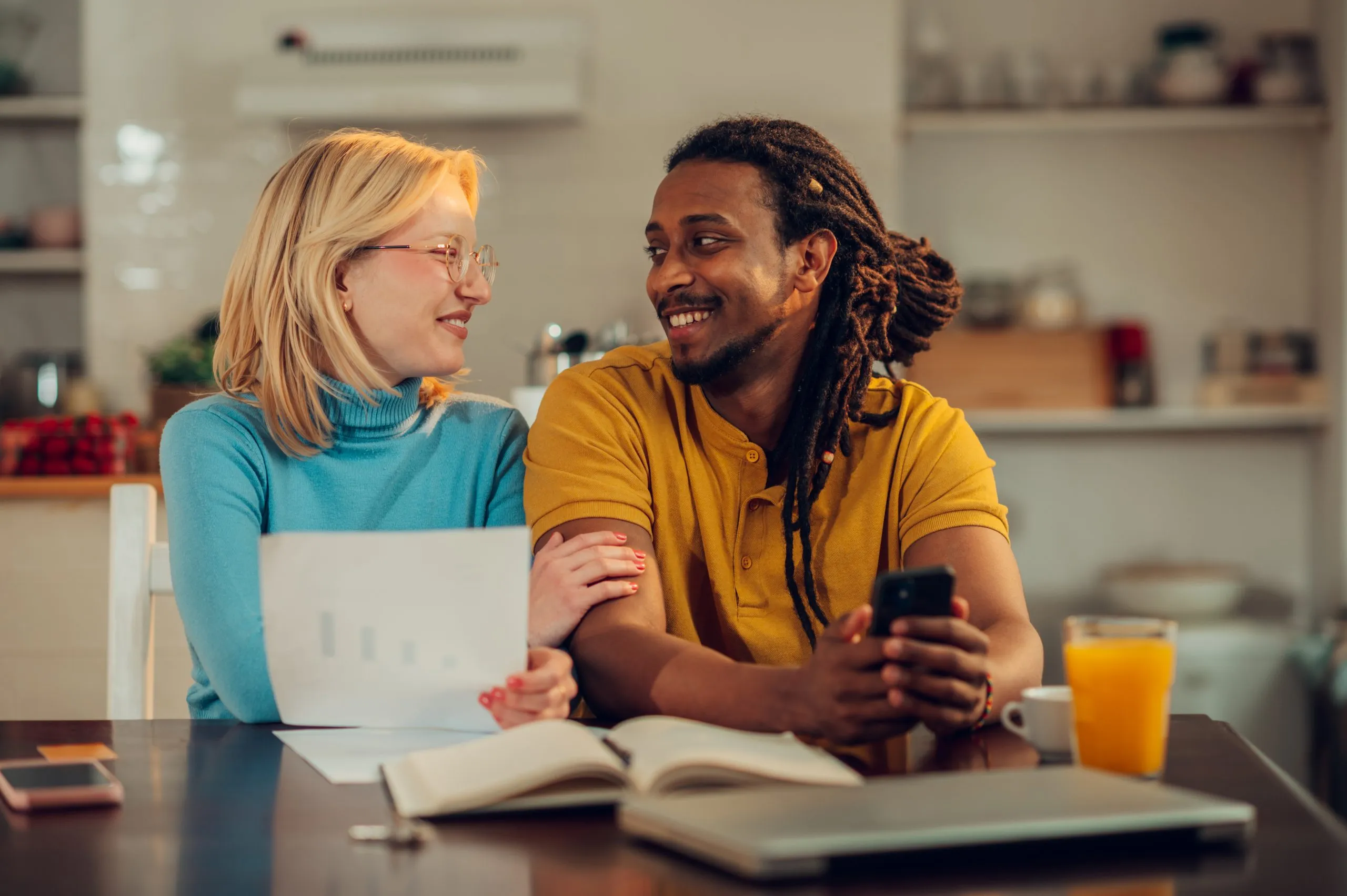 A multiracial husband and wife are planning budget and smiling at each other while holding documents, paperwork, bills, and savings and calculating them on the phone.