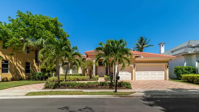 Facade of elegant colonial-style mansion in Boca Raton, with tropical front garden, cobblestone driveway, palms, trees, short grass, sidewalk, blue sky.