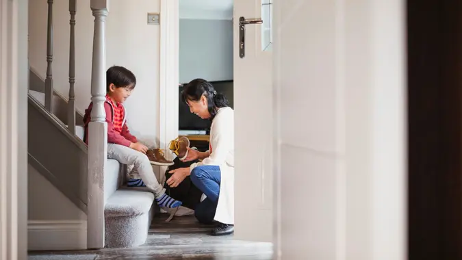 Little boy is sitting at the bottom of the stairs in his home to put his shoes on with help from his grandmother.