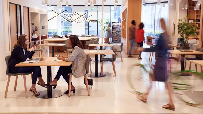 Interior Of Modern Open Plan Office With People Working And Commuters Arriving On Bikes.
