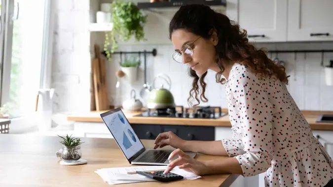 Serious young woman wearing glasses calculating finances, household expenses, confident businesswoman working with project statistics, using laptop and calculator, standing in kitchen at home.
