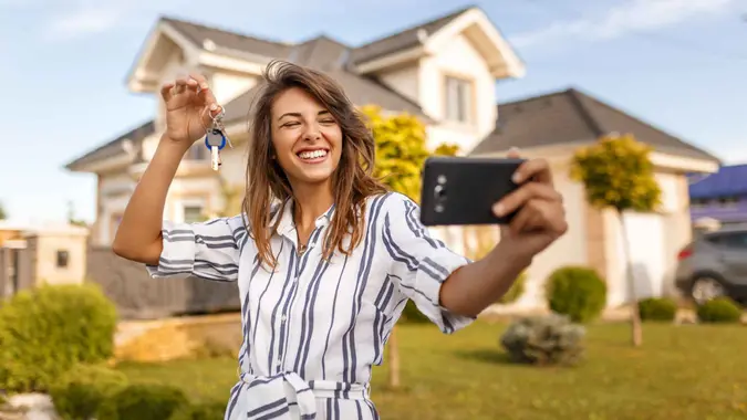 Beautiful young woman standing in front of her new house, taking a selfie using smart phone with the keys of her newly purchased property.