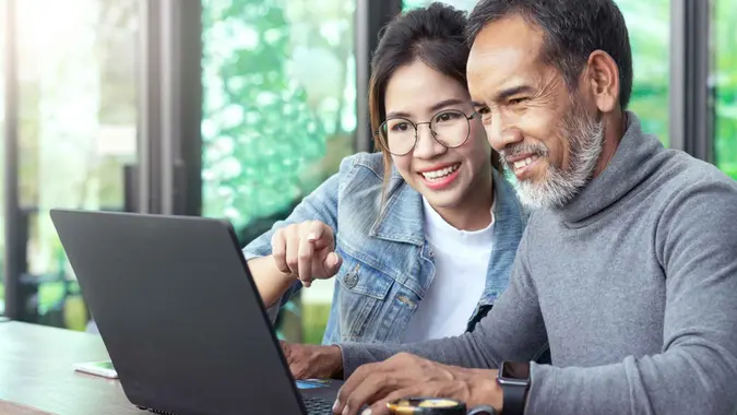 Attractive mature asian man with white stylish short beard looking at laptop computer with teenage eye glasses hipster woman in cafe.